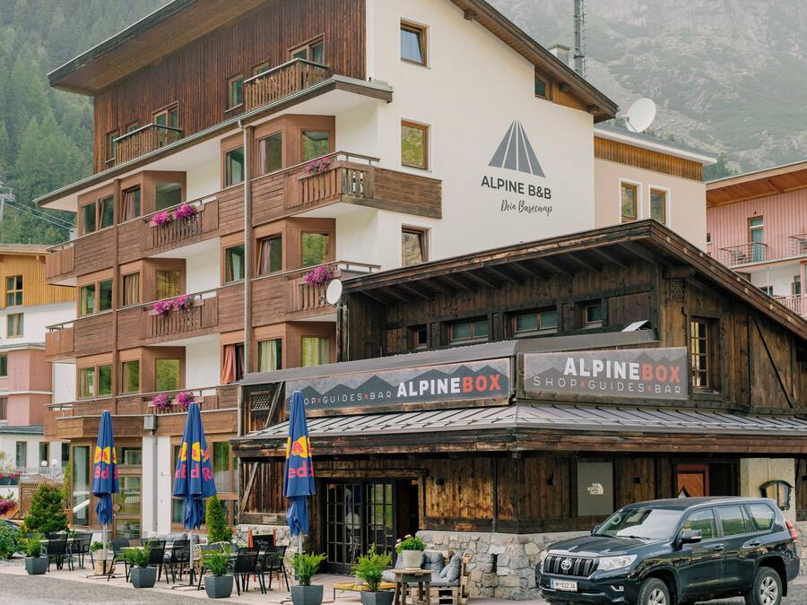 Un edificio de varios pisos con balcones, una tienda llamada "Alpine Box", mesas al aire libre con sombrillas y un coche aparcado.