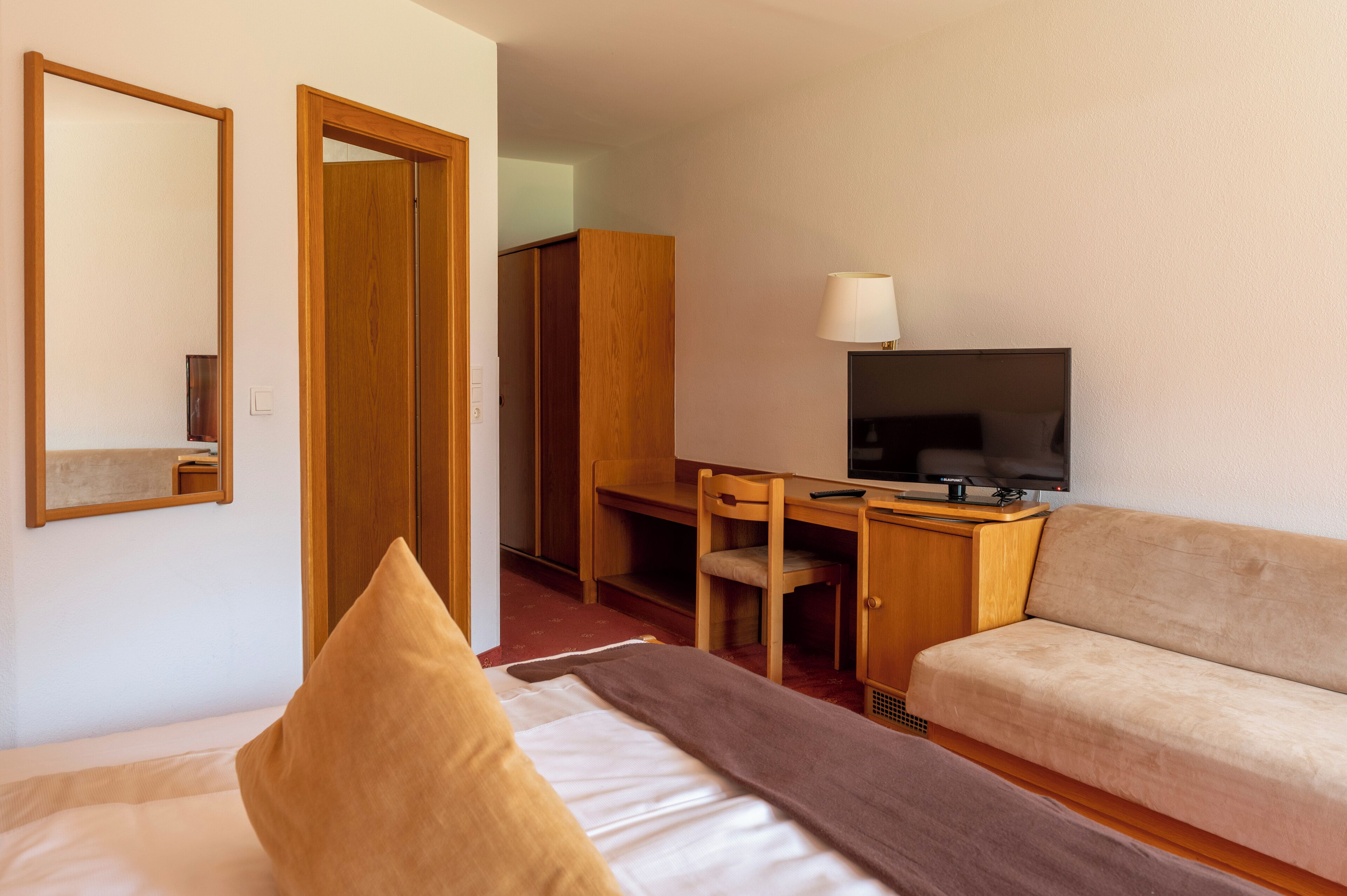 A hotel room with a bed, a sofa, a desk with a chair, a television, and a wardrobe.