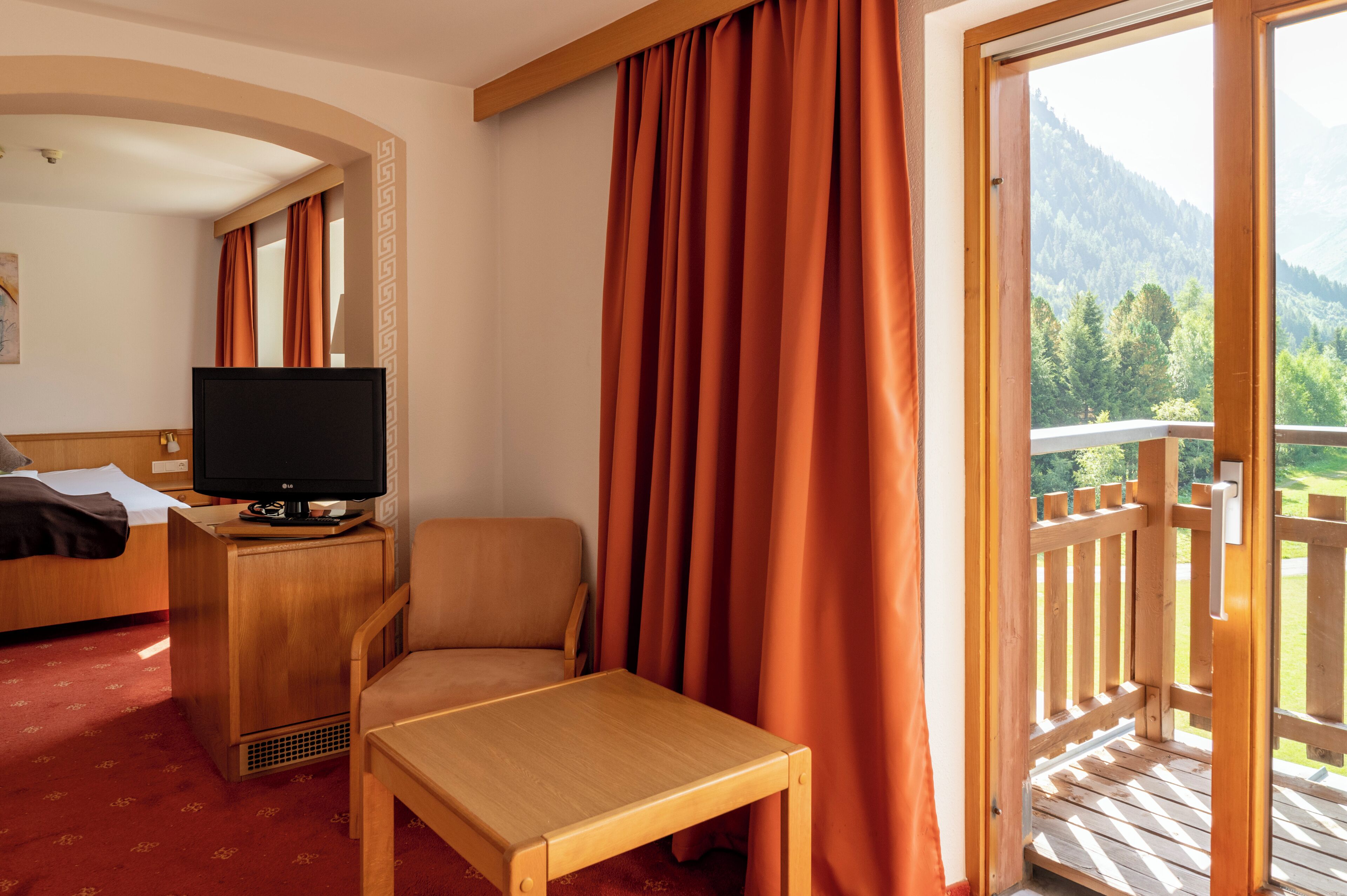 A hotel room with a bed, a desk with a television, a chair, a small table, and a balcony with a wooden railing.