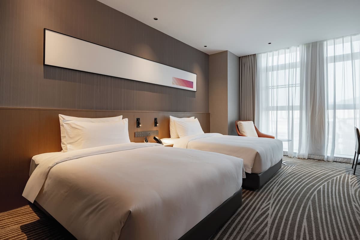 Ein Hotelzimmer mit zwei Betten, einem großen Fenster mit transparenten Vorhängen, einem Wandbild und einem modernen Kopfteil.