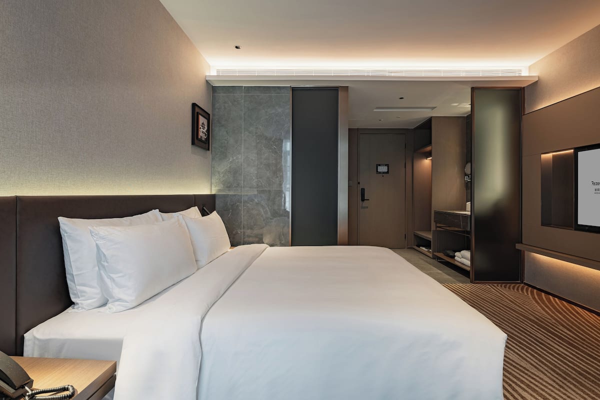 Ein modernes Hotelzimmer mit einem großen Bett, einem Nachttisch mit Telefon, einem Fernseher und einer eingebauten Ablagefläche mit Handtüchern.