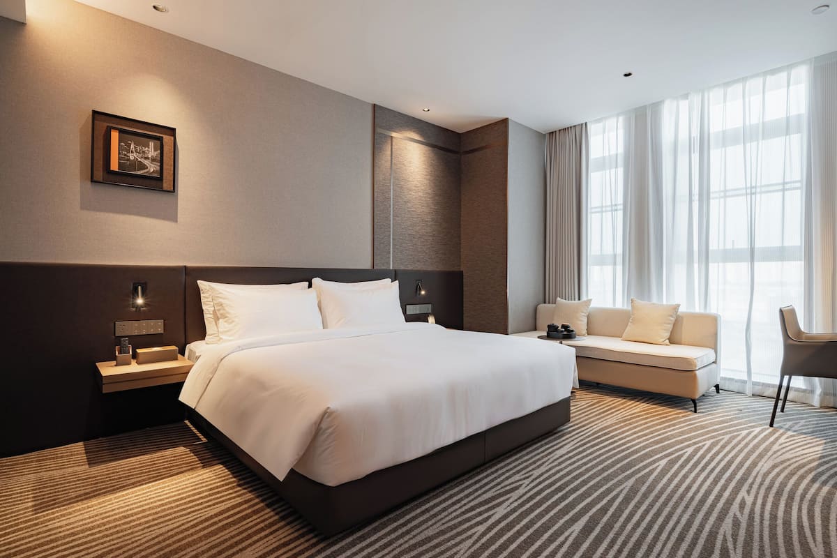 Ein modernes Hotelzimmer mit einem großen Bett, einem Sofa, einem Schreibtisch und einem großen Fenster mit Vorhängen.