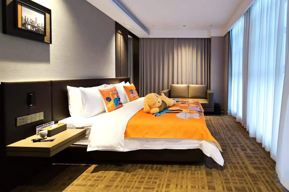 Ein Hotelzimmer mit einem großen Bett, einem Nachttisch mit Lampe, einem Sofa und einem großen Fenster mit Vorhängen.