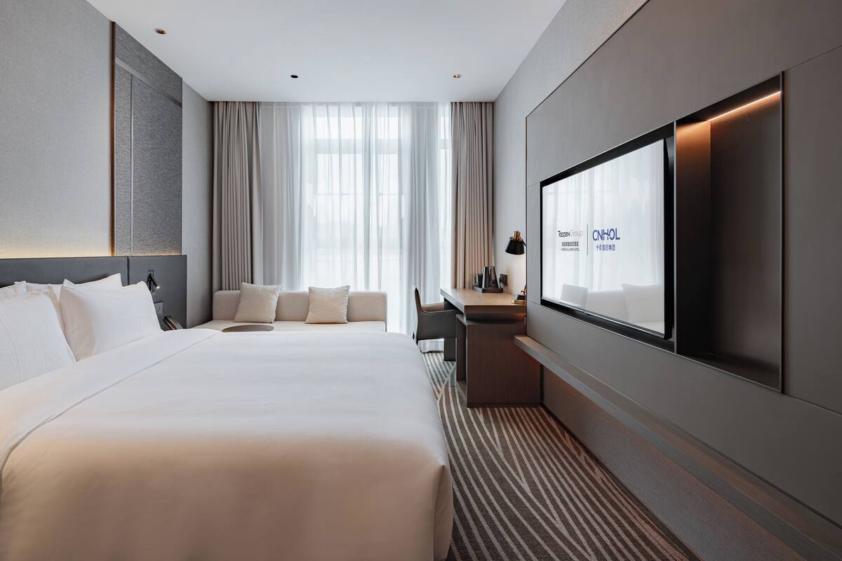 Ein modernes Hotelzimmer mit einem großen Bett, einem Schreibtisch und einem Fernsehbildschirm, das die Hotelmarke zeigt.