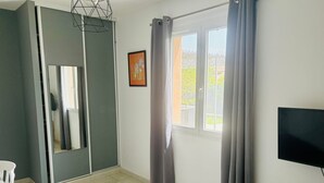 Une chambre à coucher avec un lit, une télévision, une armoire, une fenêtre avec des rideaux et un tableau au mur.
