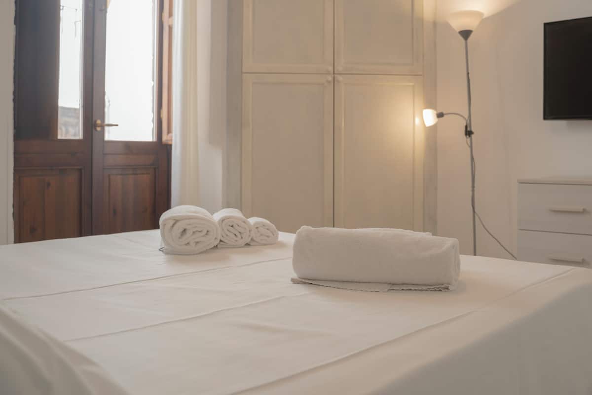A neatly made bed with rolled towels, a wardrobe, a bedside lamp, and a TV mounted on the wall.