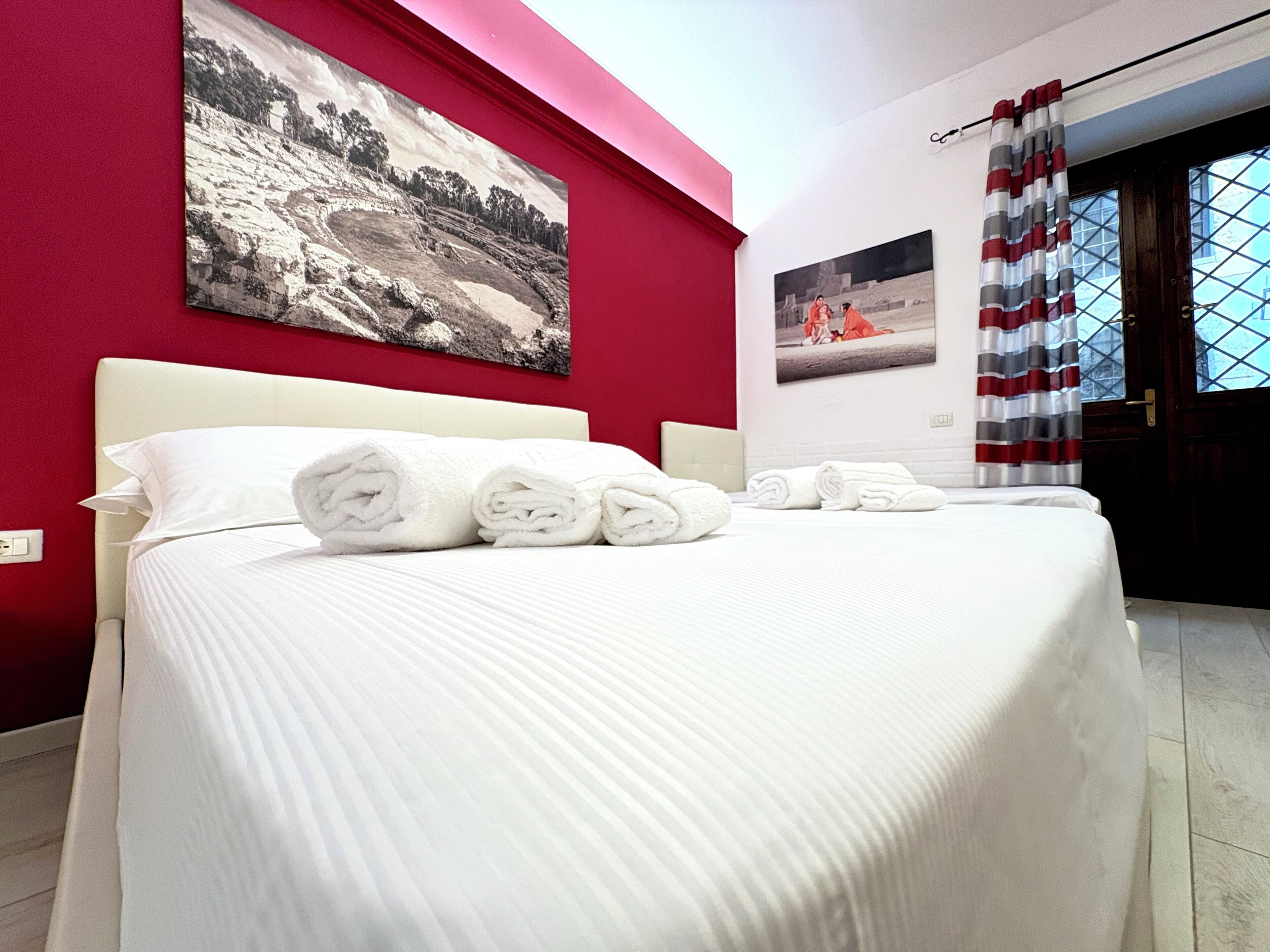 A neatly made bed with rolled white towels, a large landscape painting on the wall, and a window with curtains.