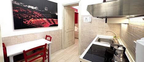 A compact kitchen with a white countertop, a built-in stove, and a sink. A dining table with red chairs is positioned under a large black and white photograph.