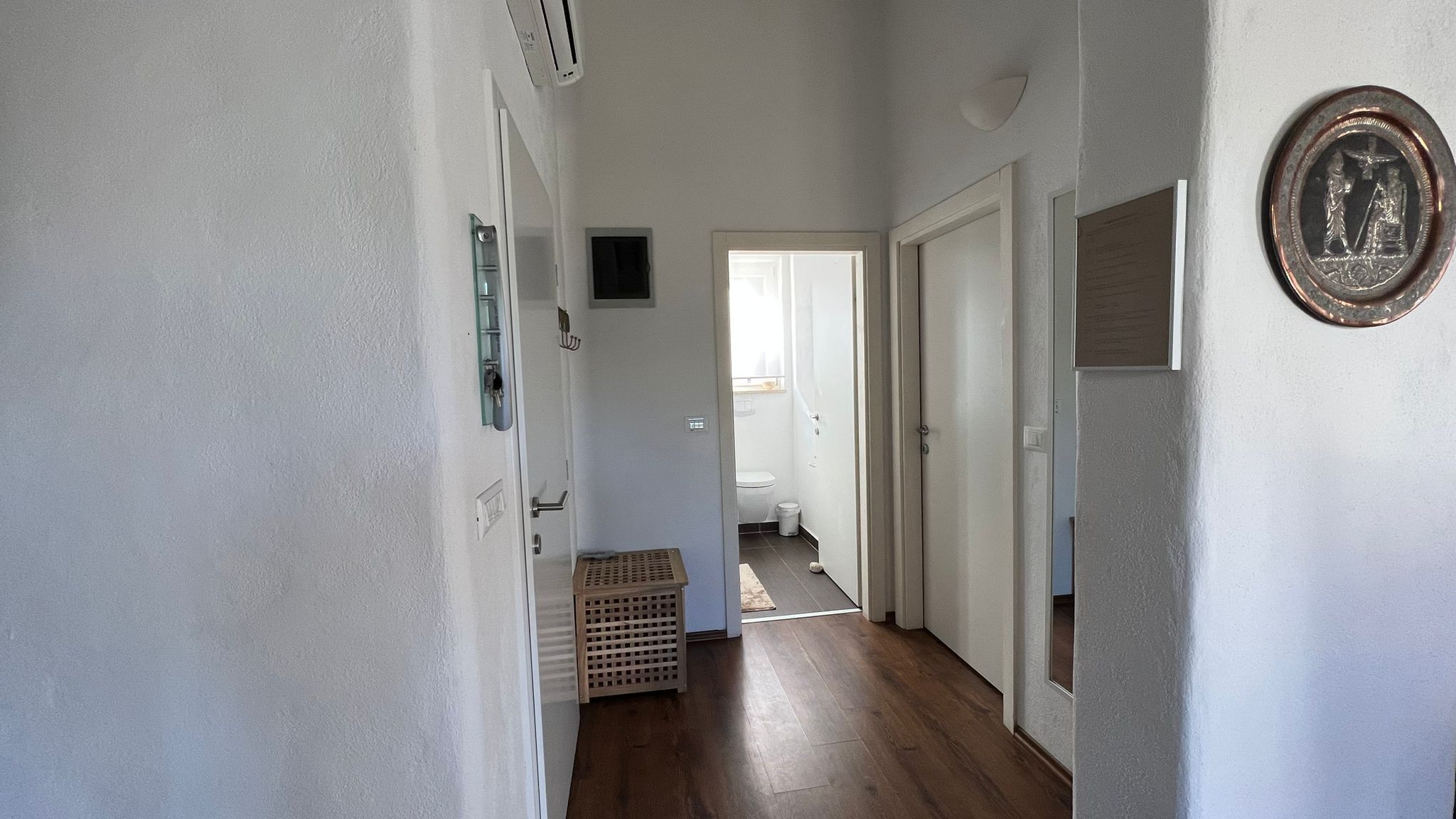 A hallway with wooden flooring, white walls, and a door leading to a bathroom.