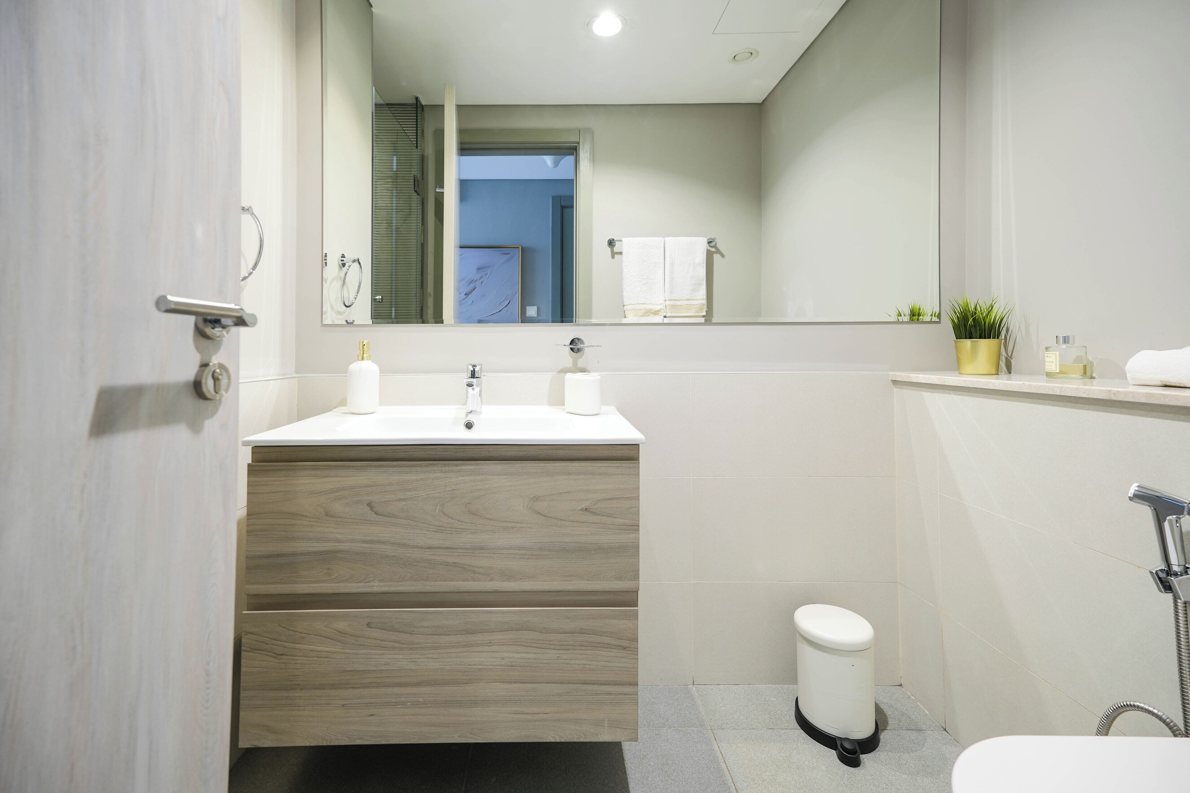 Een moderne badkamer met een houten wastafelmeubel, een witte wastafel, een spiegel, een handdoekhouder, een kleine prullenbak en een douchehoek.