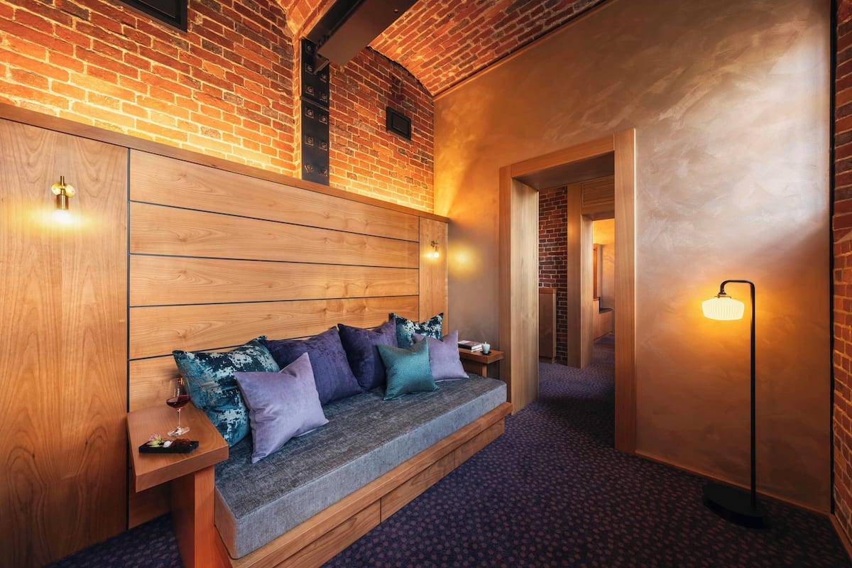A cozy room with a wooden headboard, a cushioned bench, and a small table with a glass of wine.