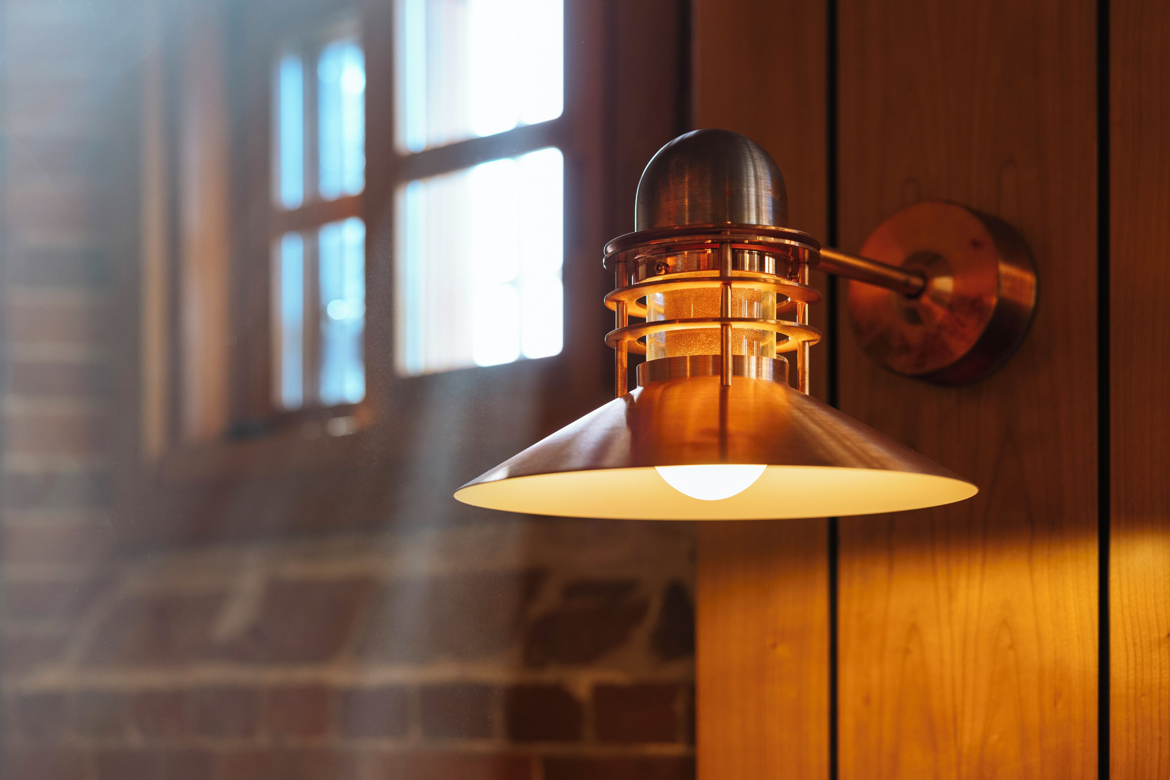 A wall-mounted lamp with a metallic finish and a conical shade, set against a wooden wall with a window in the background.
