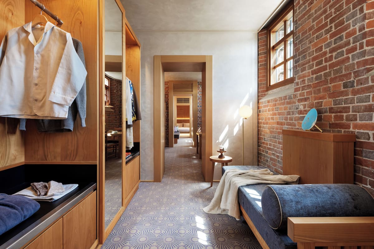 A narrow hallway with a bed, a small table with a lamp, and a brick wall.