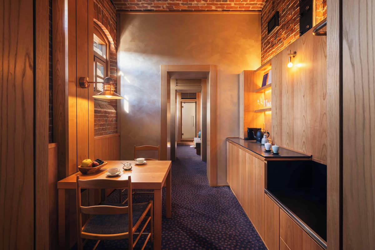 A narrow hallway with a wooden table and chairs, a bowl of fruit, and a kitchen area with a black countertop and wooden cabinets.