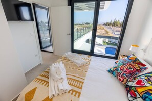 Ein Hotelzimmer mit einem Bett, einem farbenfrohen Kopfkissen, einem ordentlich gefalteten Handtuch und Blick auf einen Pool sowie Gebäude durch große Fenster.