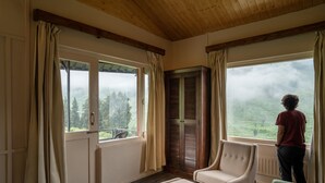 A room with a wooden ceiling, large windows with curtains, a chair, and a view of a forested landscape. - Zostel Homes Rajgundha (Multhan)