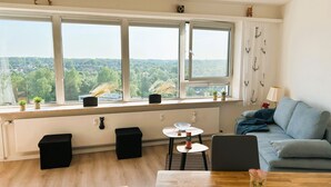 A modern living room with a large window, a blue sofa, a dining table, and a small kitchen area.