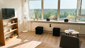 A room with a TV on a shelf, a fan, a small table with a bowl, and a window with a view of greenery.