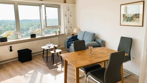 A modern living room with a wooden dining table, two chairs, a grey sofa, and a view of greenery through large windows.
