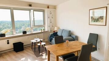 Ein modernes Wohnzimmer mit einem Holzesstisch, zwei Stühlen, einem grauen Sofa und Blick ins Grüne durch große Fenster.