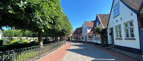 En brostensbelagt gade kantet af traditionelle europæiske bygninger, et metalhegn og frodige grønne træer.