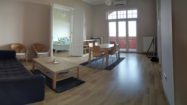 A spacious living area with a sofa, a dining table with chairs, a small kitchen area, and a balcony with a view.