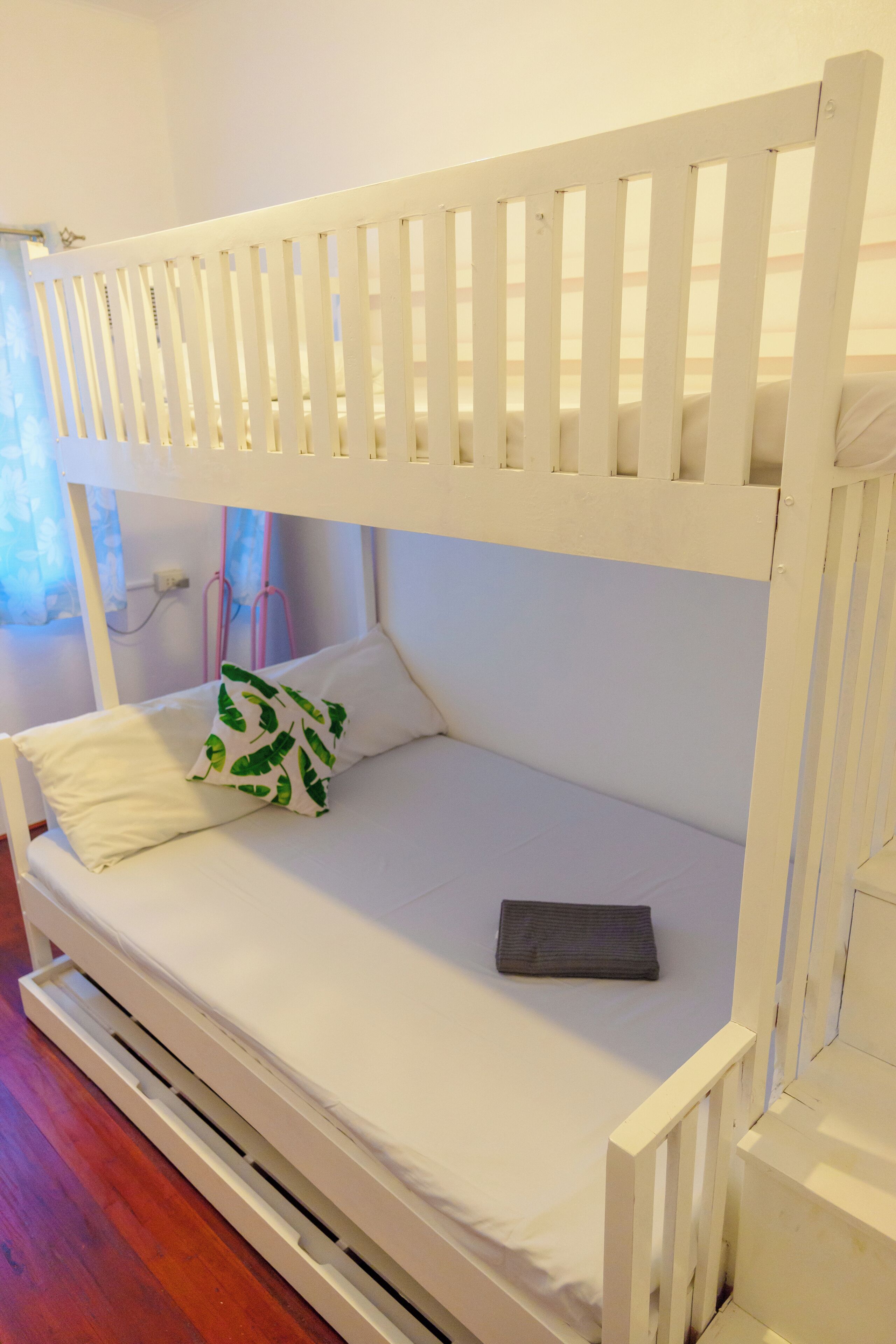Een wit stapelbed met matras en kussens, een klein nachtkastje en een raam met gordijnen.