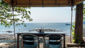 A covered outdoor seating area with a table and chairs facing a rocky beach and the sea. - Silang Beach Resort and Diving (Dauin)