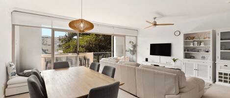 Una sala de estar moderna con comedor, ventanas grandes, ventilador de techo y televisor en la pared.