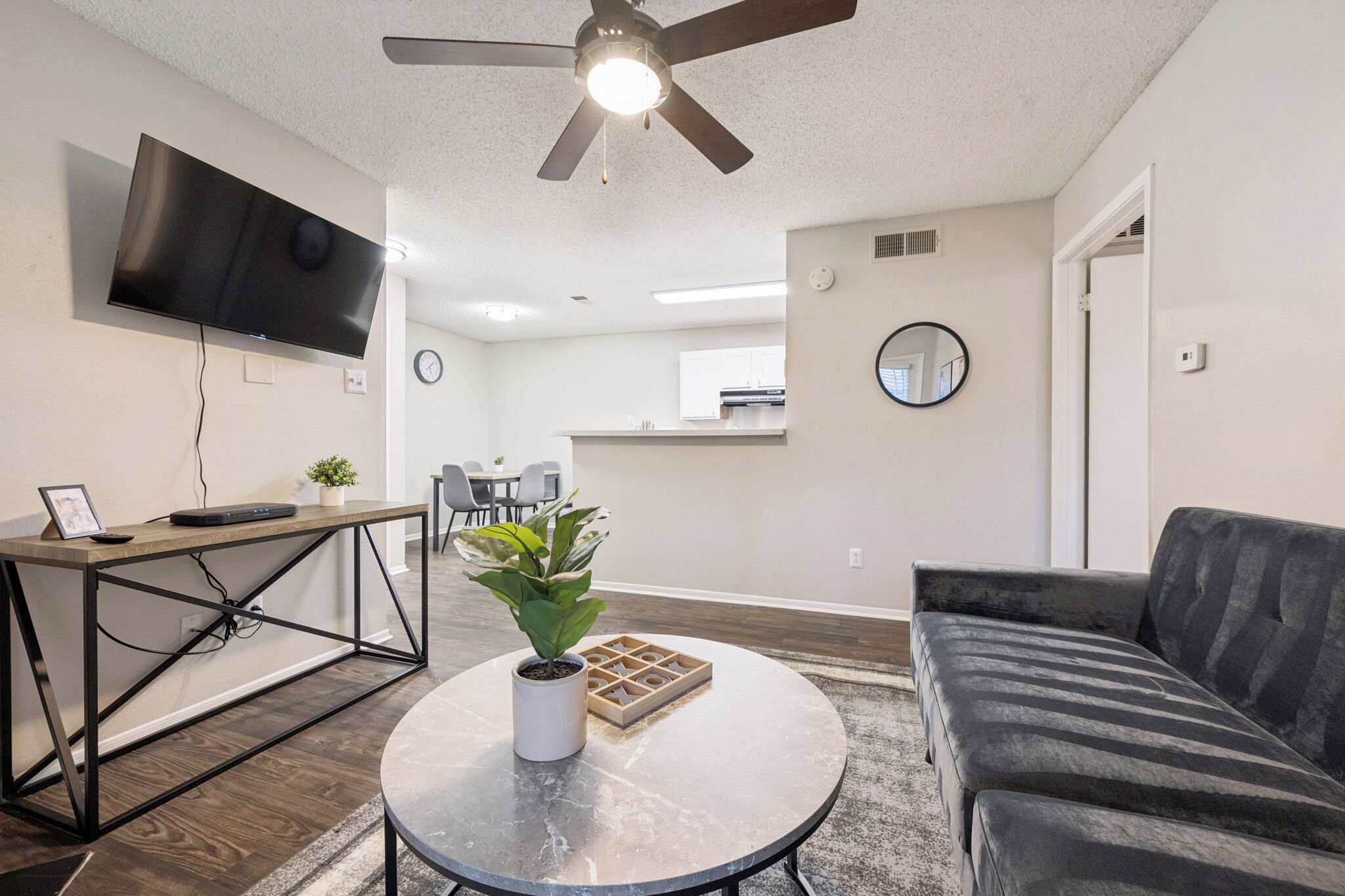 A modern living room with a flat-screen TV mounted on the wall, a ceiling fan, a round coffee table with a potted plant, and a dark-colored sofa.