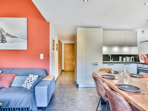 A modern living room with a sofa, a dining table set for two, and a kitchen with cabinets and appliances.