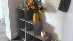 Una esquina de una habitación con un estante gris que sostiene un jarrón con flores, una taza blanca y un calentador fijado a la pared.
