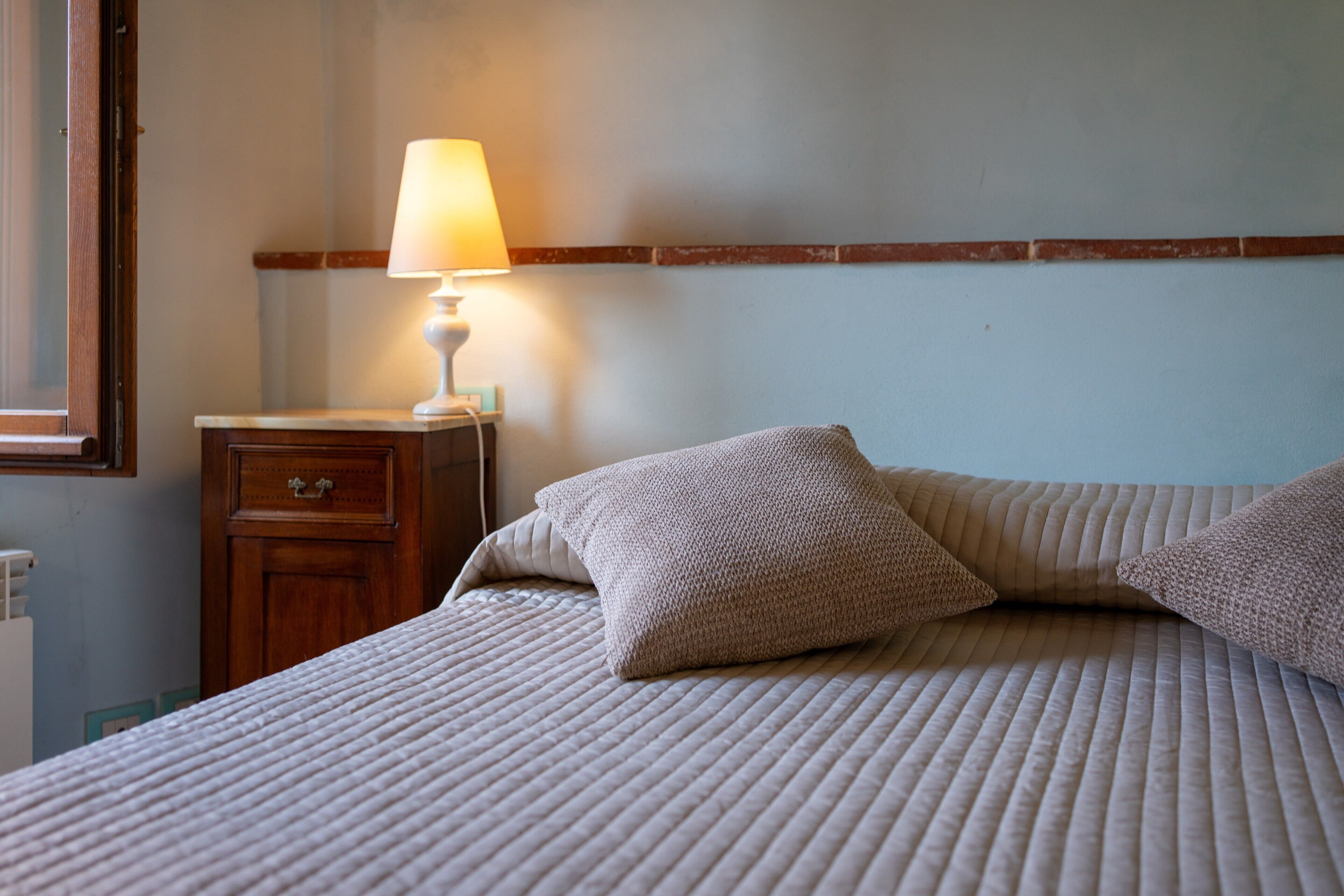 A neatly made bed with pillows, a bedside table with a lamp, and a wooden nightstand.