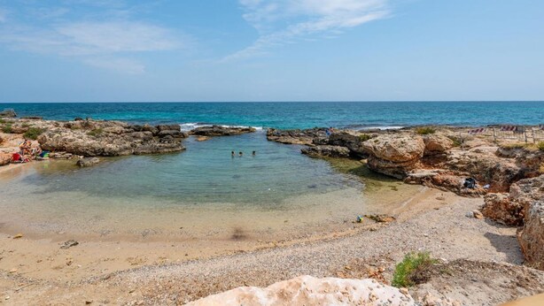 Ein felsiger Strand mit klarem Wasser, einem kleinen Becken und badenden Personen.