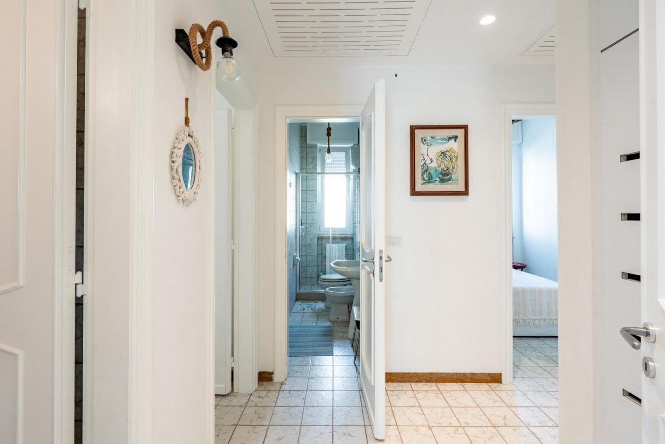 A hallway leading to a bathroom with a toilet, sink, and shower. There are doors on either side, a light fixture, and a framed picture on the wall.
