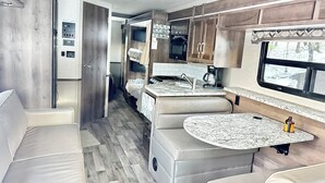 Une cuisine compacte pour camping-car, dotée d’une cuisinière intégrée, d’un évier et d’une fenêtre donnant sur la forêt.