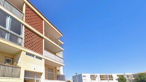 A multi-story residential building with balconies, a clear sky, and a parking area in the foreground.