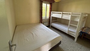 A small, single-room accommodation with a bunk bed, a single bed, a window with curtains, and a ceiling light.