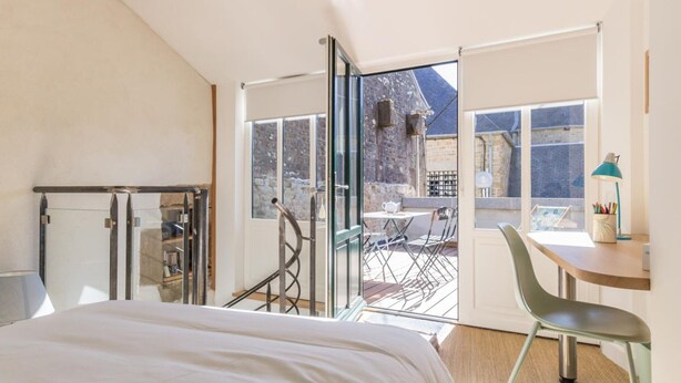 A bedroom with a bed, a desk, and a chair, a balcony with a table and chairs, and a view of a brick building. - Charming duplex with private terrace in the heart of Avranches (Avranches)