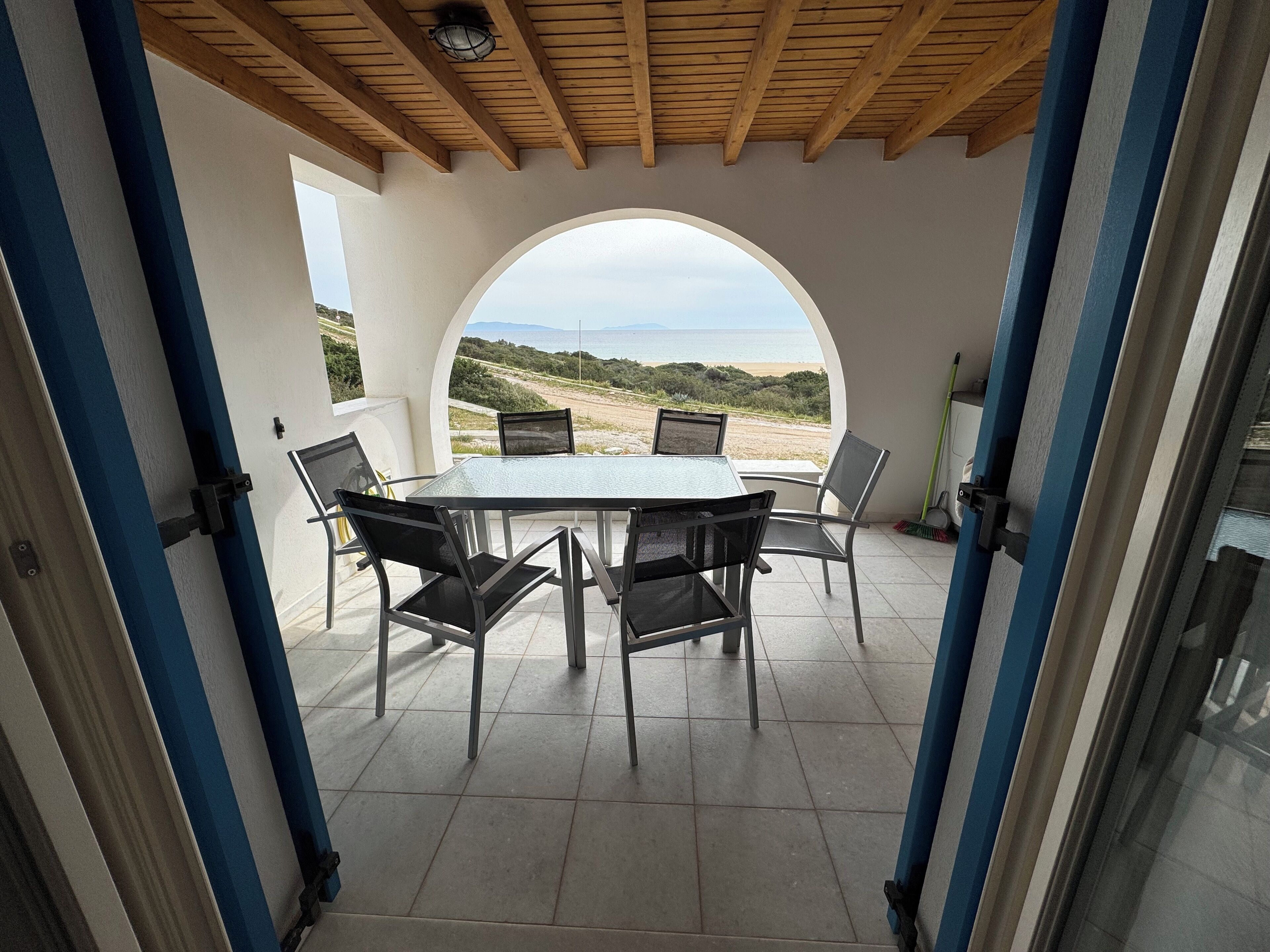 A covered patio with a table and chairs, a view of the beach, and a wooden ceiling.