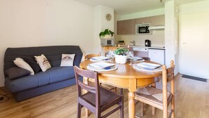 A modern living room with a dark blue sofa, wooden dining table, and chairs. There is a kitchen in the background with a microwave, stove, and cabinets.