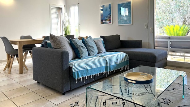 A modern living room with a grey sofa, a glass coffee table, and a dining area with a wooden table and chairs.