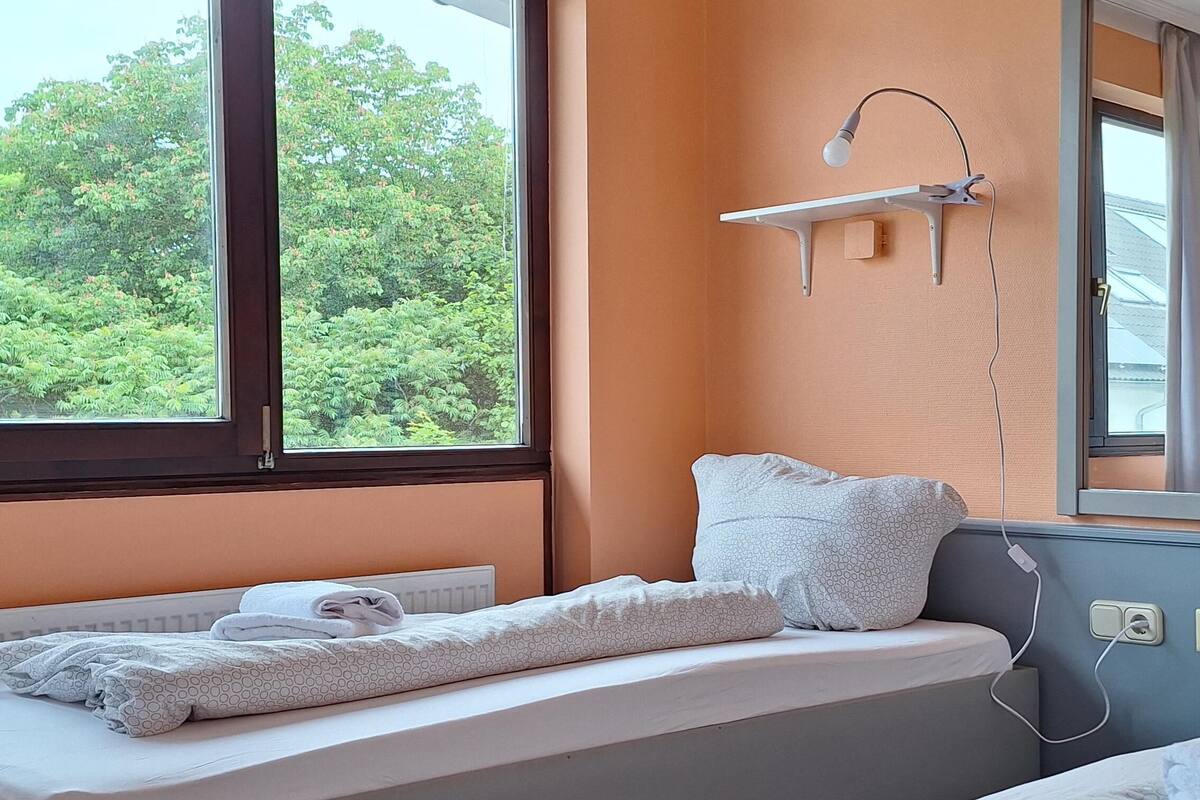 Un lit bien fait, avec une couette blanche et des oreillers, une table de chevet avec une lampe, et une vue sur la verdure à l’extérieur.