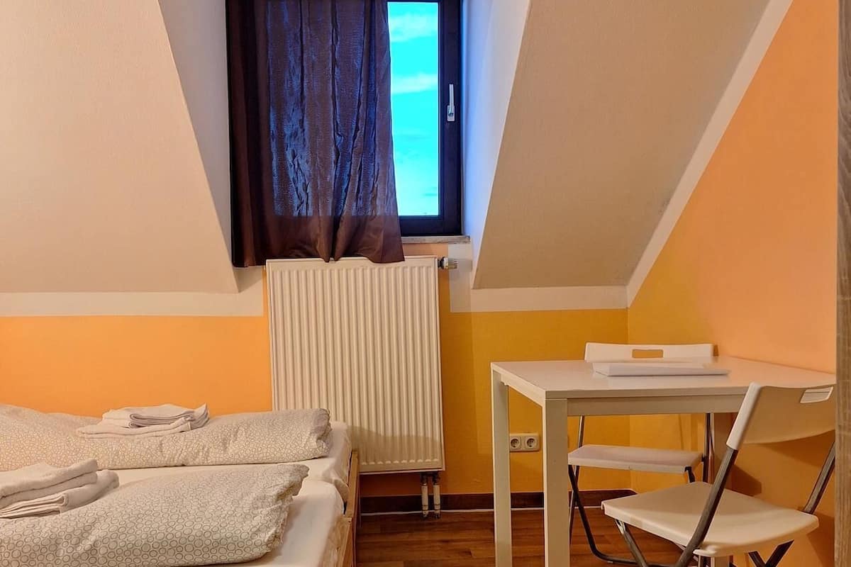 Une petite pièce agréable et confortable, avec un lit, une table et une chaise. Il y a une fenêtre avec des rideaux, un radiateur et une vue sur le ciel.