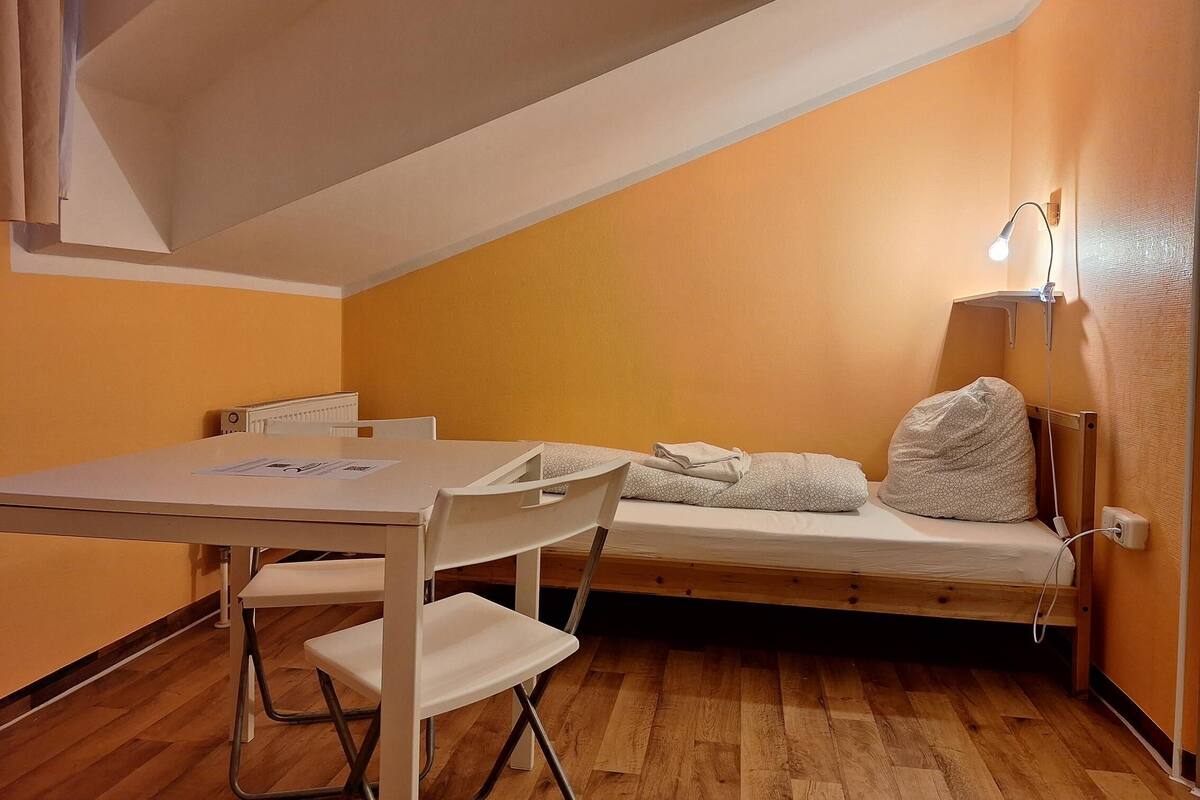 Une petite pièce agréable et chaleureuse, avec un plancher en bois, un lit doté d’un matelas et de coussins blancs, une petite table avec des chaises et une lampe fixée au mur.
