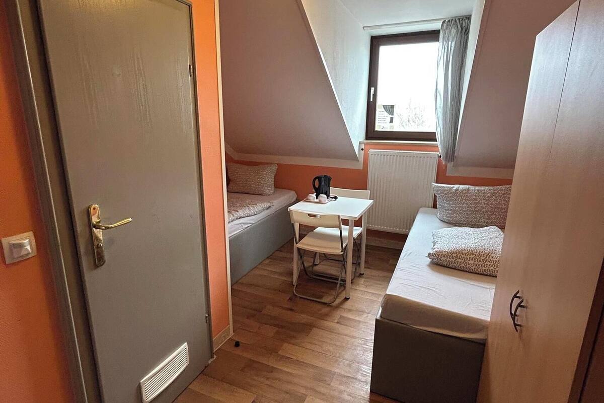Une petite pièce comprenant un lit, une petite table et une chaise. Il y a une porte à gauche et une fenêtre à droite.