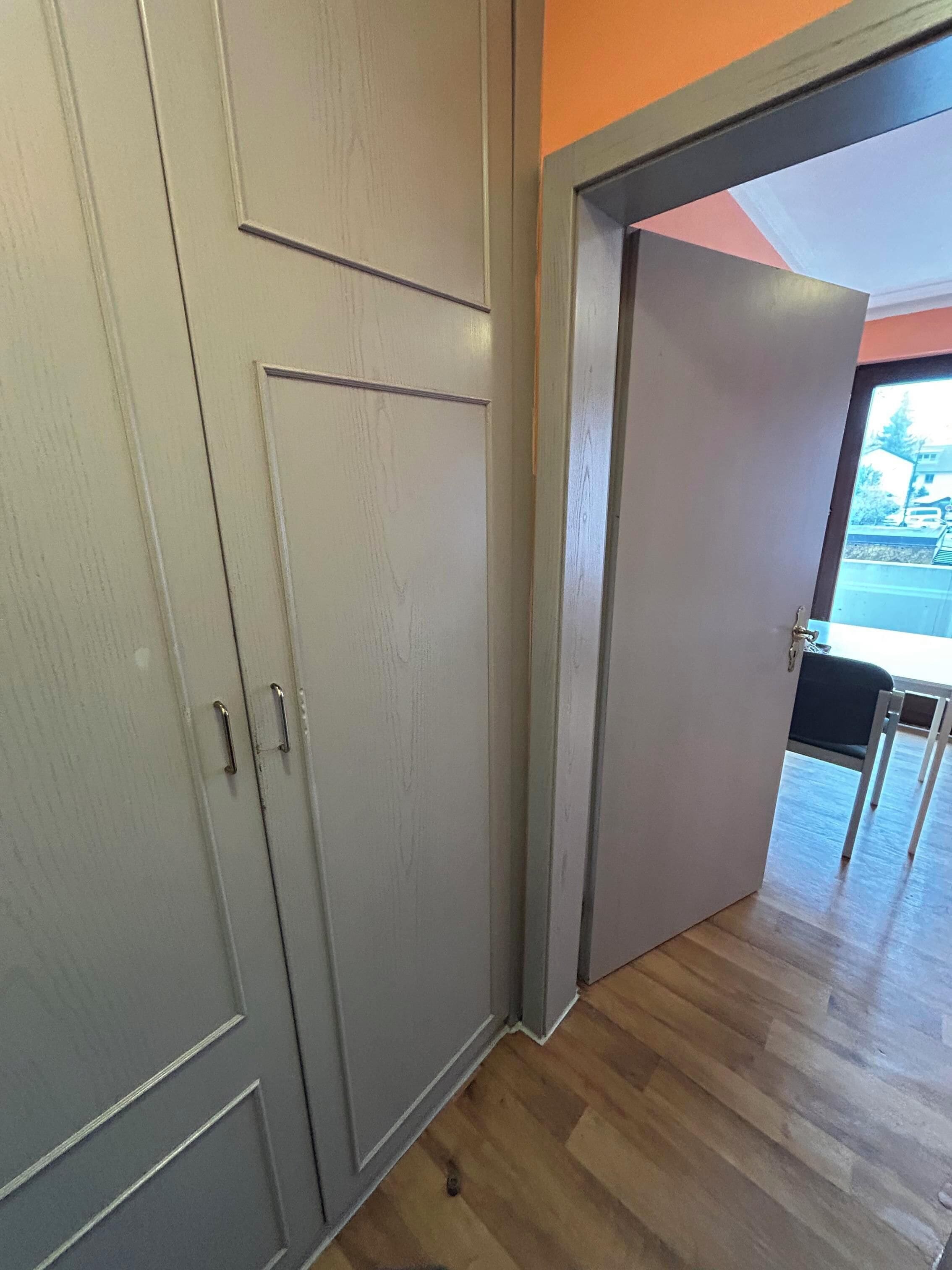 Un pasillo con un armario gris, suelo de madera y una puerta abierta que da a una habitación con vistas a la calle.