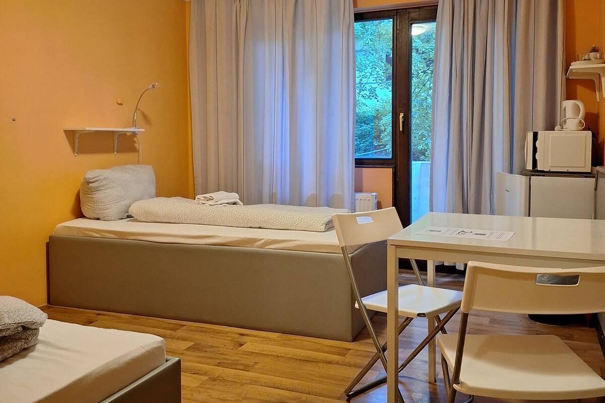 Une chambre d’hôtel de taille réduite, comprenant un lit, un petit bureau et une chaise. On y trouve une fenêtre avec des rideaux, un radiateur et un luminaire au plafond.