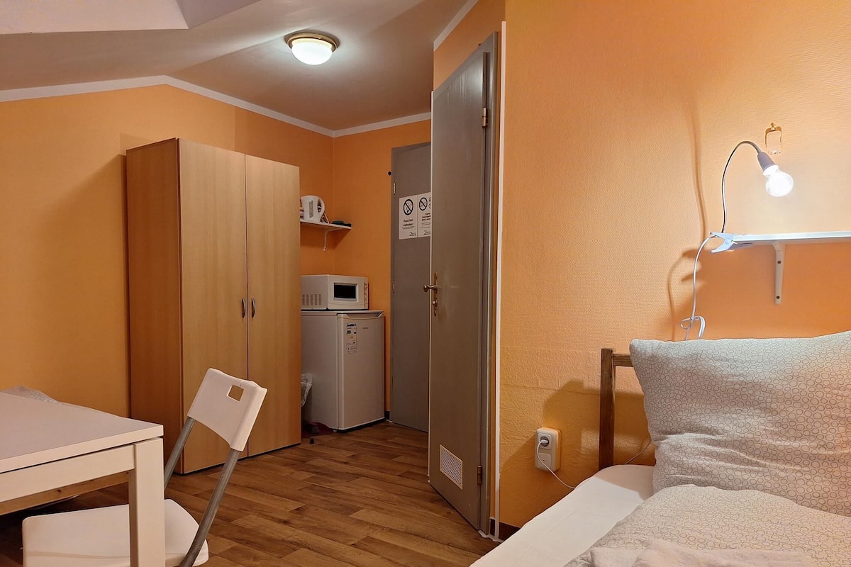 Une chambre compacte comprenant un lit, une table de chevet et un bureau avec une chaise. On peut également apercevoir une armoire et une petite kitchenette équipée d’un micro-ondes et d’un réfrigérateur.