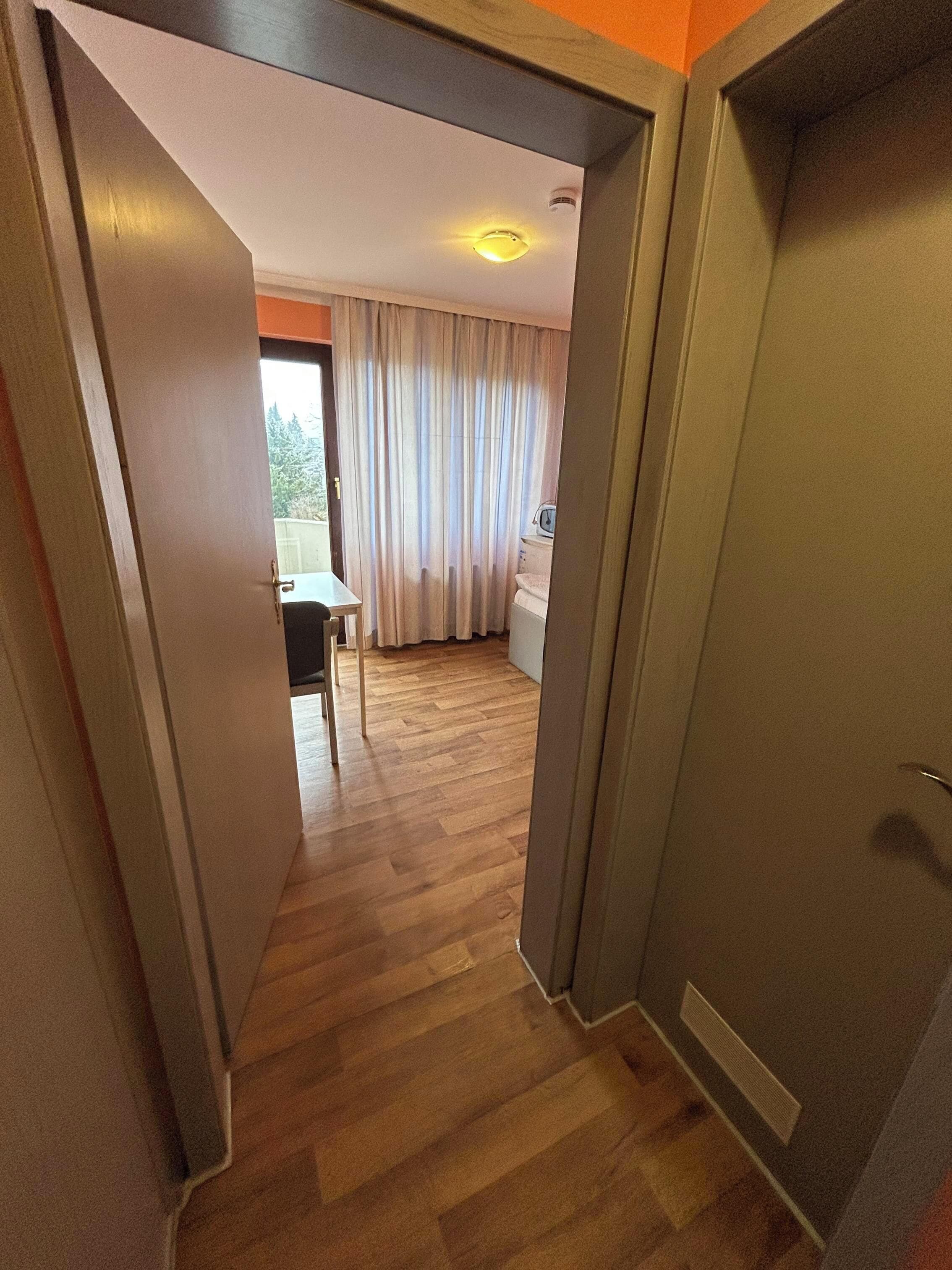 A hallway with wooden flooring leading to a room with a bed, a window with curtains, and a visible bathroom.